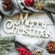 Merry Christmas in gold writing with a grey blanket border decorated with snow-dusted fir branches, holly berries, pine cones, baubles and gold snowflake shapes.